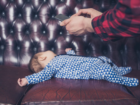 The hands of a man using smartphone to take photo of a sleeping baby on a leather sofa