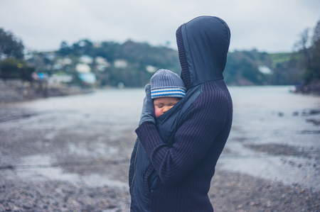A young mother is standing outside by a river at low tide with her baby inside her jacketの写真素材