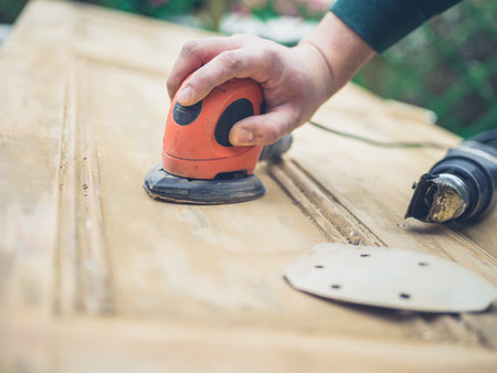 A young man is sanding an old door with an electric sander