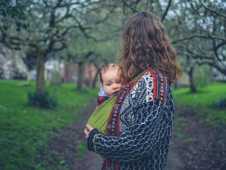 A young mother is standing in the park with her baby in a carrier slingの写真素材