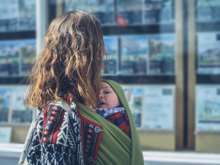 A young mother is looking at an estate agent's window with her baby in a carrier slingの写真素材