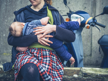 A young mother is breastfeeding her baby outside with a scooter parked behind herの写真素材