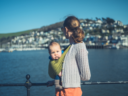 A young mother is standing by the river in a small village with her baby in a slingの写真素材