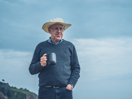 A senior man is standing on the beach with a cup of drink in his handの写真素材