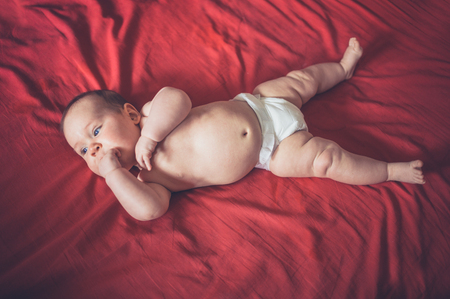 A cute little baby in his diaper is lying on a red bed sheetの写真素材