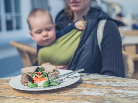 A mother with a baby in a sling is sitting at a table outside with a sandwich