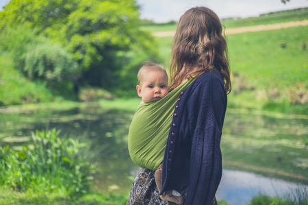 A young woman with a baby in a sling is standing by a pond in the countrysideの写真素材