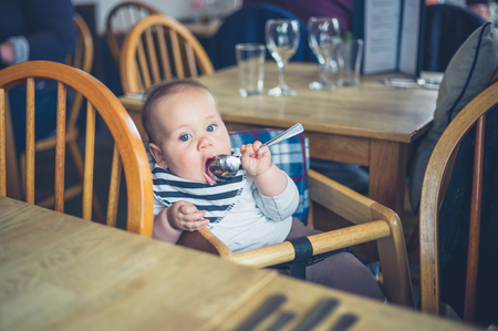 A little baby is sitting at a table in a restaurant and is eating with a spoonの写真素材