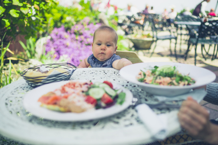 A little baby is sitting at atable outside and is looking at all the foodの写真素材