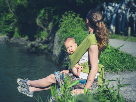 A young mother with her baby in a sling is relaxing by the riverの写真素材