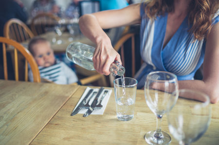 A young woman is at a table poring water with her little baby in the background