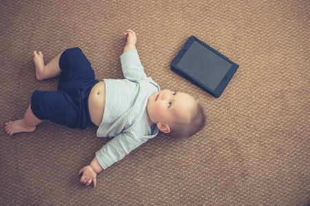 A cute little baby is lying on the floor with a tablet computerの写真素材