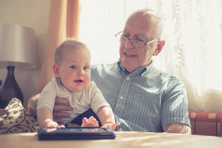 A grandfather is sitting at a table with his grandchild and is using a tabletの写真素材