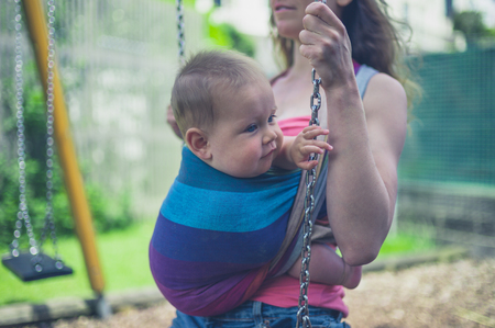 A young mother is sitting on a sling in the playground with her baby in a slingの写真素材