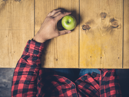 A young man is sitting at a wooden table with an apple in his handの写真素材