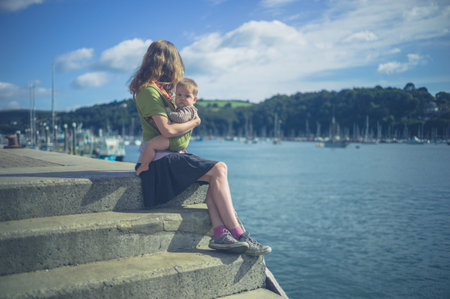 A young mother with her baby in a sling is sitting by the river in a village in summer