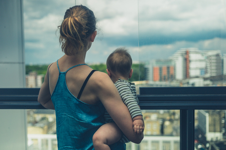 A young mother is standing on the balcony of her high rise apartment in the city holding her babyの写真素材