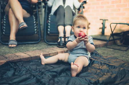 A cute little baby is sitting on the ground in the garden and is eating a peachの写真素材