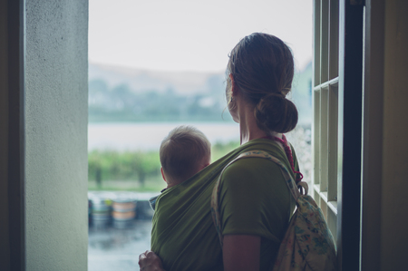A young mother with her baby in a sling is standing in a doorwayの写真素材