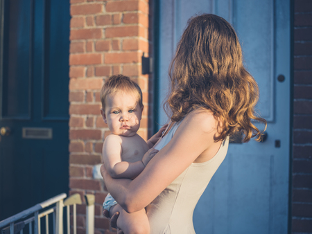 A young mother is standing outside a house at sunset with her baby in her armsの写真素材