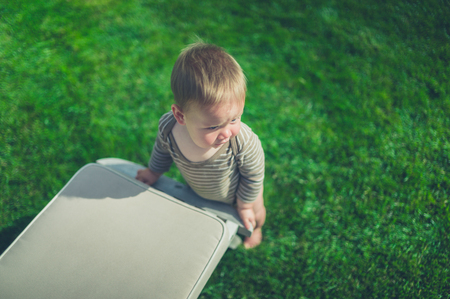 A cute little baby is using a deck chair in the garden to help him stand upの写真素材
