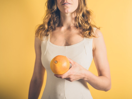 A young woman is standing against a yellow background holding a grapefruitの写真素材