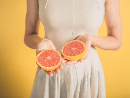 A young woman against a yellow background is holding two grapefruit halvesの写真素材