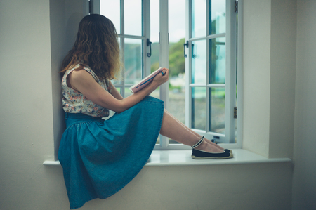 A young woman is sitting and reading by the window of a country houseの写真素材