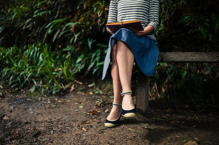 A young woman is sitting on a bench in the forest and is reading a bookの写真素材