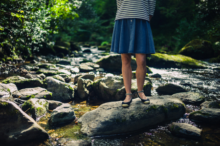 A young woman wearing a skirt is standing on some rocks in the river on a summer dayの写真素材