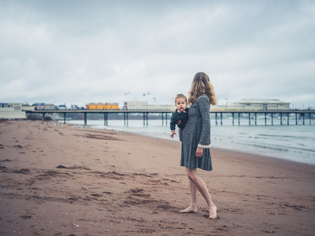 A young mother is standing with her baby on the beach looking at a pierの写真素材