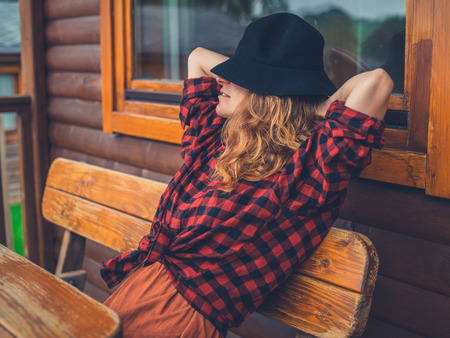 A young woman wearing a hat is relaxing on the porch of a log cabinの写真素材