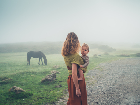 A young mother with her baby in a sling is standing in the fog looking at a horseの写真素材