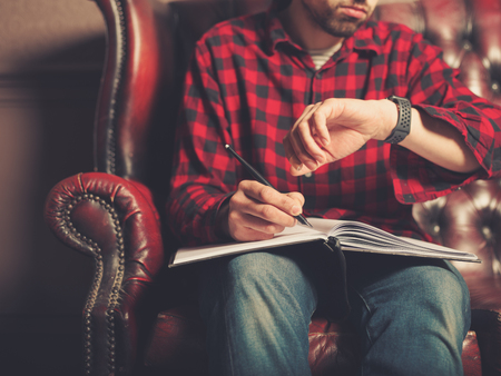A young man wearing a smart watch is sitting on a leather sofa and is writing in a notebookの写真素材