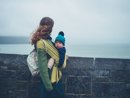 A young mother with her baby in a sling is standing by the seaside on a cloudy dayの写真素材