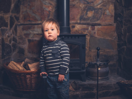 A toddler is standing by a log burner holding a shovelの写真素材