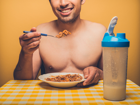 A young fitness man against yellow backdrop is eating mince and protein shakeの写真素材