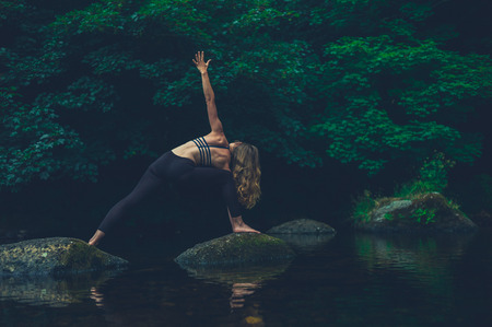 A young woman doing yoga on a rock in the river is in a warrior poseの写真素材