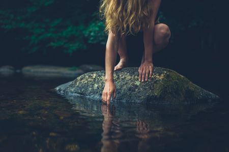 A young woman is sitting on a rock in the river and is touching the waterの写真素材