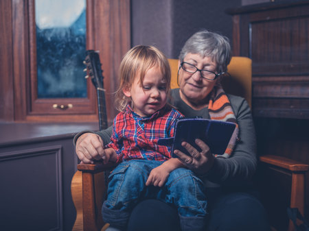 A little toddler and his grandmother are sitting at home looking at a smartphone