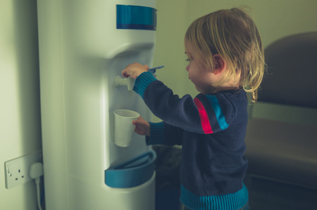 A little toddler is getting a glass of water from the water coolerの写真素材