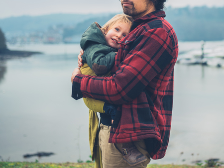 A young father with his toddler in a sling is by a river with boatsの写真素材