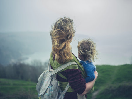A young mother with her toddler in a sling is standing on top of a hill by the sea on a windy day