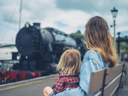 A young mother and her toddler are sitting on a bench at the station watching a steam train arrive