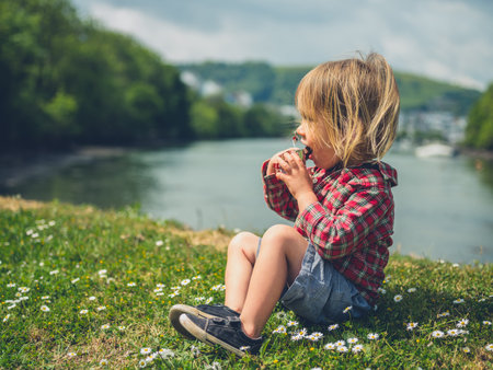 A little toddler is sitting on the grass by the water eating a fruit bar