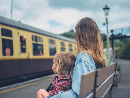 A young mother and her toddler are sitting on a bench at the station watching a train arriveの写真素材