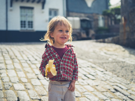 A little toddler is eating a banana outdoors in a cobbled street