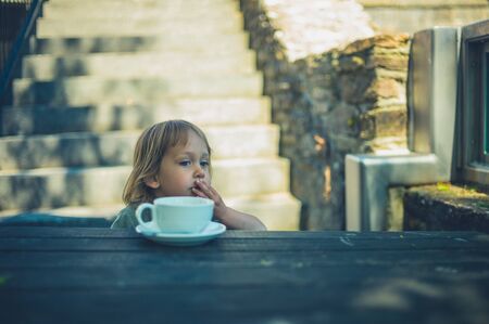 A little todler is sitting at a table with a coffee cup in front of him outdoorsの写真素材