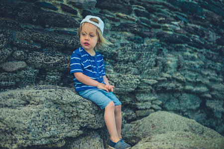 A little toddler is sitting by a stone wall in a small town on a summer day