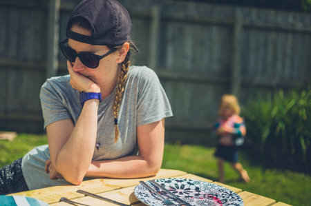 A young woman is sitting at a table outdoors with a little toddler in the background